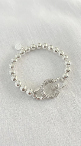 A silver beaded bracelet with a circular charm featuring a stylized letter G, displayed on a textured white fabric background.