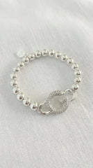 A silver beaded bracelet with a circular charm featuring a stylized letter G, displayed on a textured white fabric background.