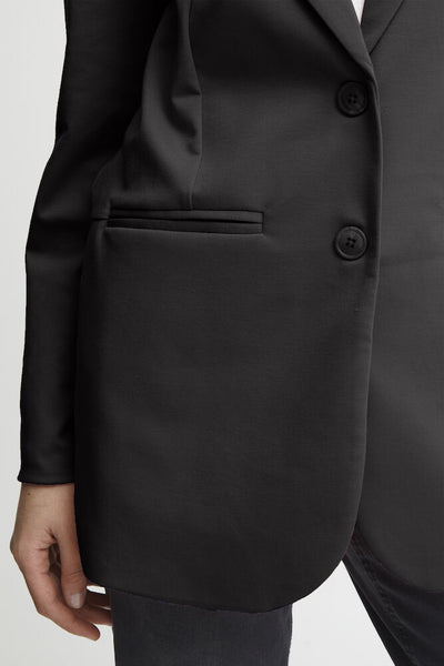 Close-up of a black blazer jacket with two dark buttons, a welt pocket, and a hand visible at the side, against a light neutral background.