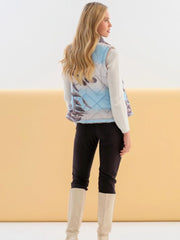 A woman shown from the back wearing a sleeveless quilted vest with a blue, gray, and brown abstract pattern, a white long-sleeve knit top, black pants, and beige knee-high boots, set against a split white and beige studio background.