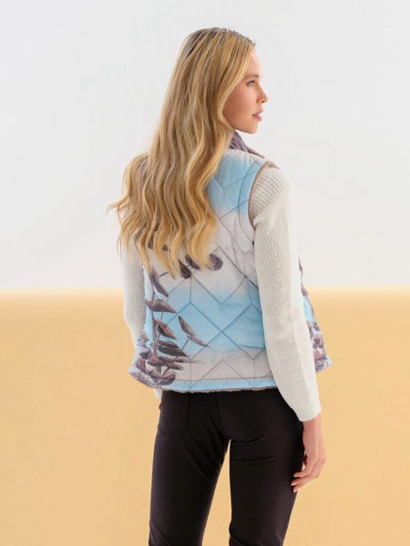 A woman shown from the back wearing a sleeveless quilted vest with a light blue and gray argyle pattern over a white ribbed sweater and black pants, against a split white and beige studio background.