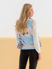 A woman shown from the back wearing a sleeveless quilted vest with a light blue and gray argyle pattern over a white ribbed sweater and black pants, against a split white and beige studio background.