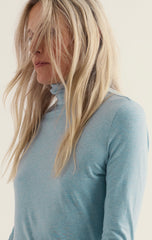 A close-up side profile of a woman with long blonde hair wearing a light blue  long-sleeve turtleneck top against a plain off-white background.