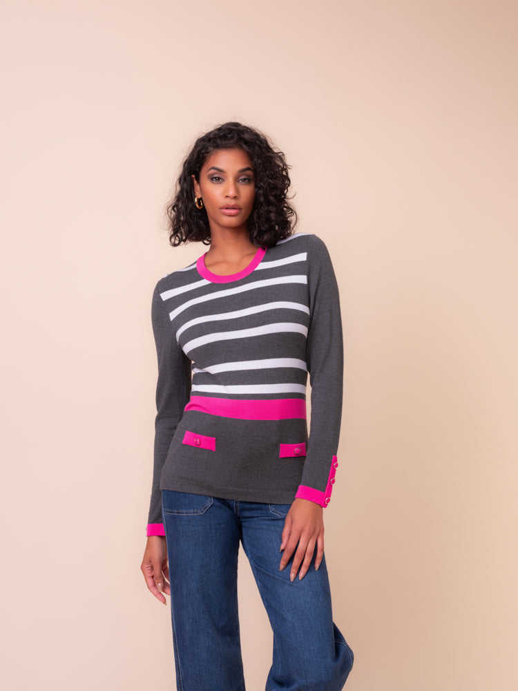 A woman modeling a dark grey long-sleeve knit sweater with white horizontal stripes, bright pink trim, and matching pink pocket details, paired with blue jeans against a solid beige background.