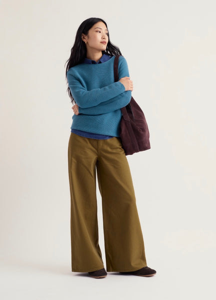 A woman stands against a plain light background wearing Seasalt blue knit sweater over a blue collared shirt, olive wide-leg pants, and dark shoes, while carrying a dark plum tote bag.
