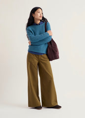 A woman stands against a plain light background wearing Seasalt blue knit sweater over a blue collared shirt, olive wide-leg pants, and dark shoes, while carrying a dark plum tote bag.