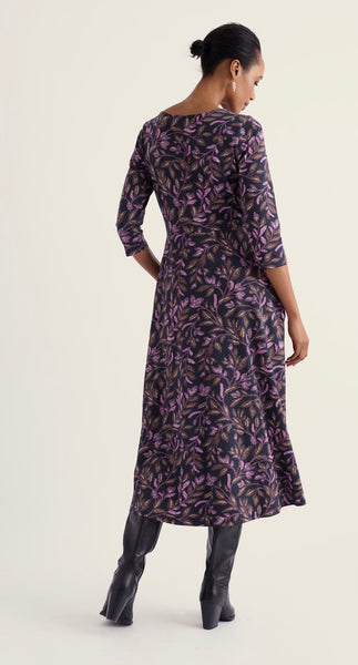 A woman shown from the back wearing Seasalt knee-length dress with three-quarter sleeves and a dark purple and black leafy floral pattern, paired with black knee-high boots, against a light beige studio background.