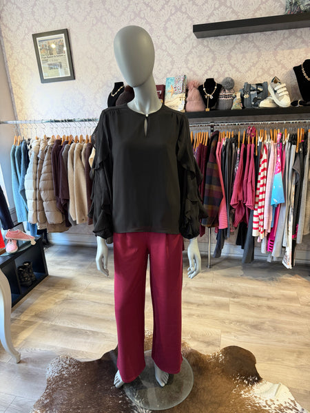 A mannequin in a clothing boutique wearing a black long-sleeve blouse with ruffled sleeves and a split neckline, paired with bright magenta wide-leg pants. Clothing racks and shelves are visible in the background.