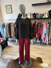 A mannequin in a clothing boutique wearing a black long-sleeve blouse with ruffled sleeves and a split neckline, paired with bright magenta wide-leg pants. Clothing racks and shelves are visible in the background.