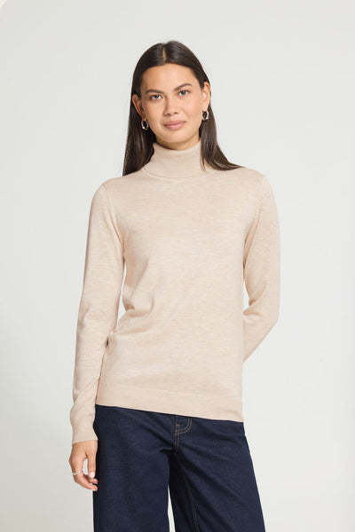A woman modelling a Ichi light beige turtleneck sweater with dark navy pants, standing against a plain light grey studio background.