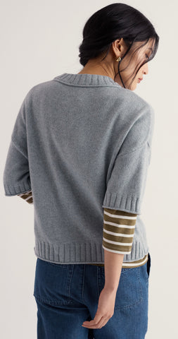 A woman modelling Seasalt light blue knit sweater with olive green and white striped elbow top, standing on a sandy beach with the ocean in the background.