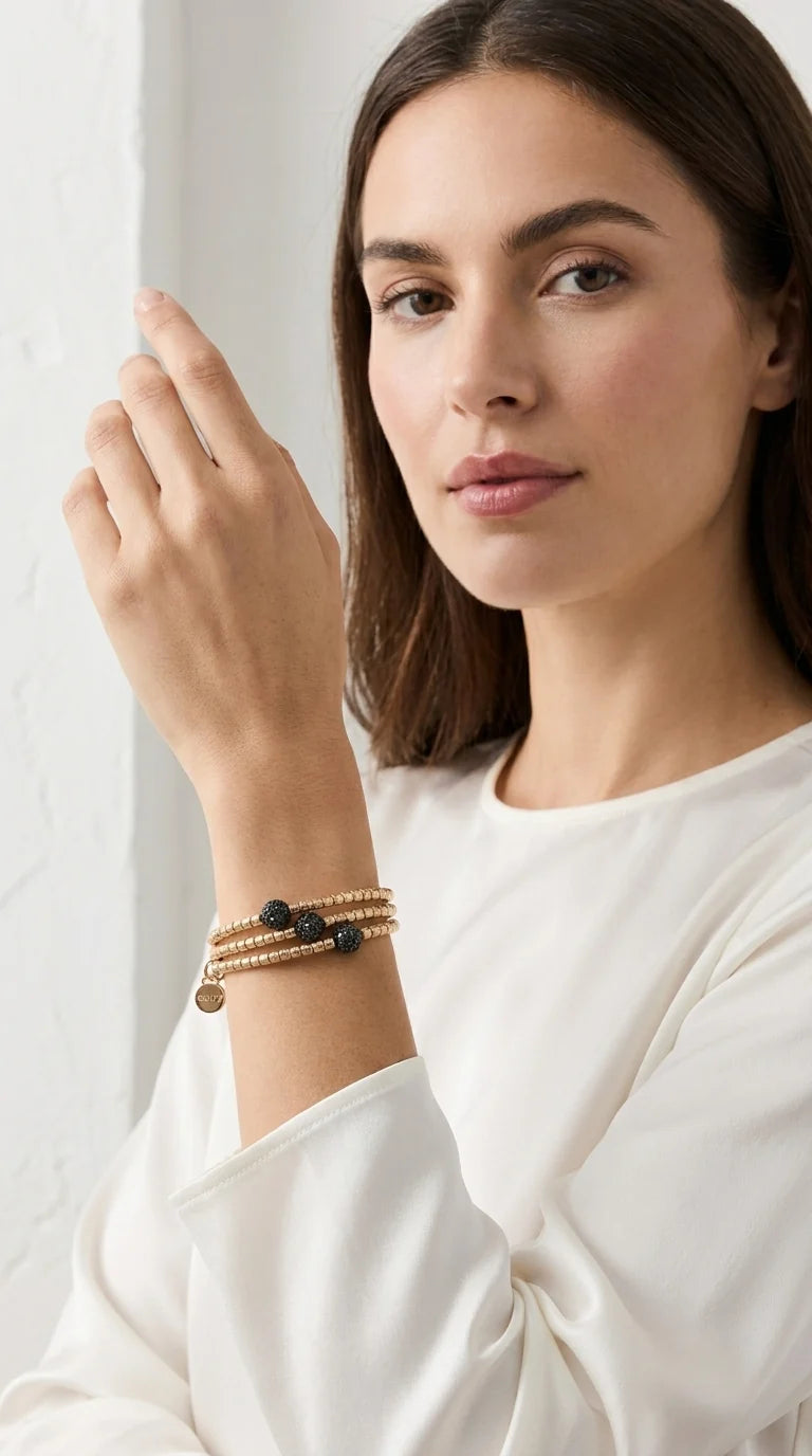 A woman wearing a delicate stack of three beaded Envy bracelets on her wrist, featuring gold-toned metal and dark beads, against a soft white background.