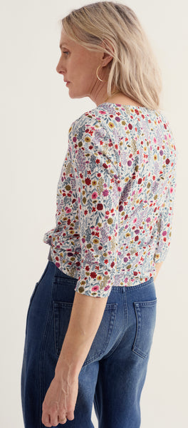 A woman shown from the back wearing Seasalt short-sleeve blouse with a dense multicolor floral print on a light background, paired with dark blue jeans, against a plain off-white studio background.