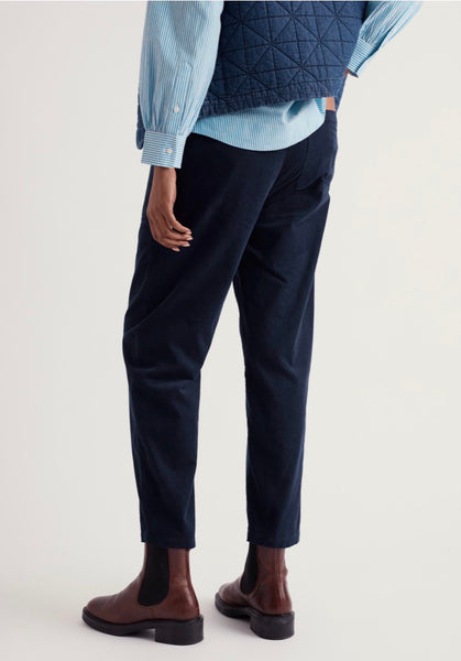 A person shown from the back wearing Seasalt dark navy trousers, a light blue striped shirt, a navy quilted vest, and brown Chelsea boots against a plain light background.