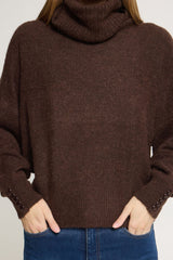 A person wearing a Ichi dark brown ribbed knit roll neck sweater with button details on the cuffs, paired with blue jeans against a light grey background.