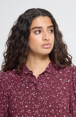 A woman  wearing an Ichi burgundy button-down shirt with a small white floral pattern, shown from the chest up against a light grey background.