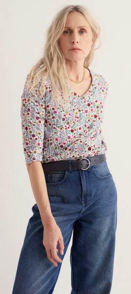 A woman modelling a Seasalt short sleeve V neck top with a dense multicolour floral and paisley print, paired with a black belt and blue jeans, against a plain light background.
