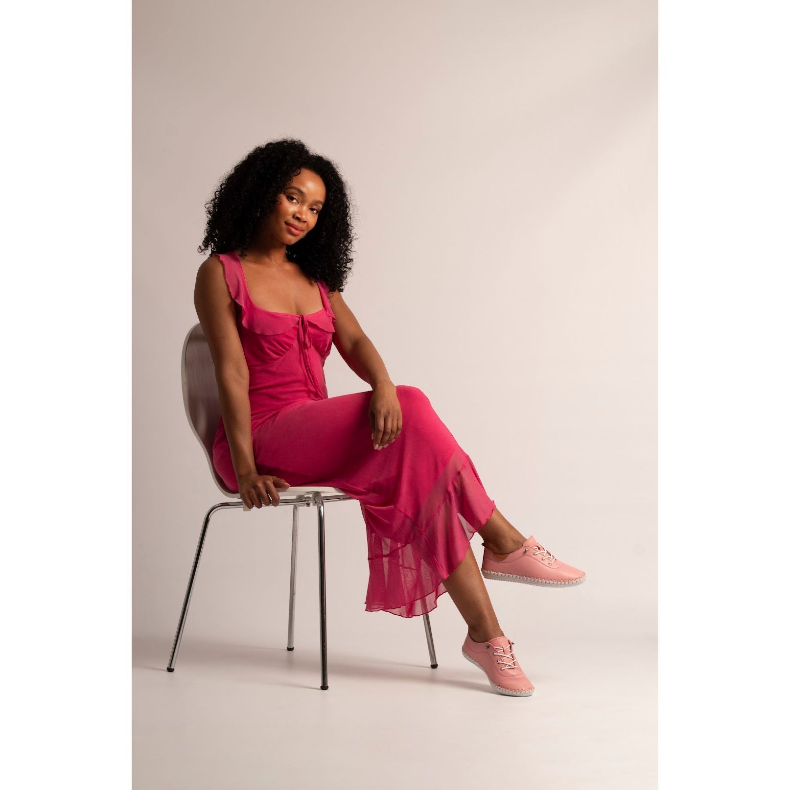 Woman in a pink dress sitting on a chair against a plain background wearing pink plimsoll.