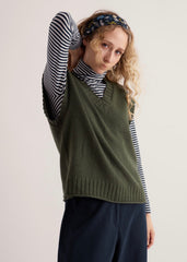 A Seasalt Vest olive top over a striped long sleeve roll neck top, wearing blue trousers. With a beige white studio background.
