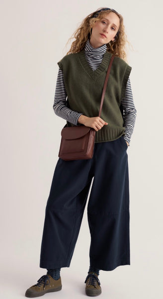 A woman models Seasalt dark olive green knit vest over a black and white striped turtleneck, paired with wide-leg navy trousers, brown shoes, and a brown leather crossbody bag, set against a plain light beige studio background.