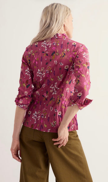 A woman shown from the back wearing a Seasalt pink floral long-sleeve blouse with pleated detailing and olive green trousers, against a plain light background.