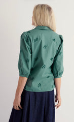 Model showing back of green embroidered 3/4 sleeve blouse worn with a blue skirt.