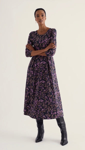 A woman modeling a Seasalt long-sleeved midi dress with a purple and black botanical leaf pattern, paired with black leather knee-high boots, against a plain off-white studio background.