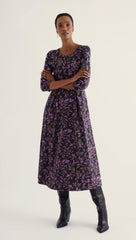 A woman modeling a Seasalt long-sleeved midi dress with a purple and black botanical leaf pattern, paired with black leather knee-high boots, against a plain off-white studio background.