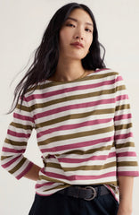 A woman wearing a Seasalt 3/4 sleeve striped top with horizontal bands of olive green, dusty pink, and off-white, paired with a black belt and dark trousers, against a plain light background.