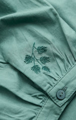 Close-up of a sage green linen shirt featuring a dark green embroidered pine branch on the chest pocket and a matching button.