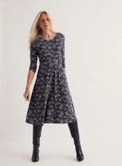 A woman modelling a Seasalt dark grey three-quarter sleeve A-line dress with a purple and green floral pattern, paired with black knee-high boots, against a plain light grey studio background.