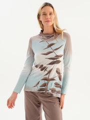 A woman modeling a long-sleeve Dolcezza cowl-neck top with a gradient design transitioning from light blue to taupe, featuring a brown botanical leaf pattern printed across the front, paired with taupe pants against a white studio background.
