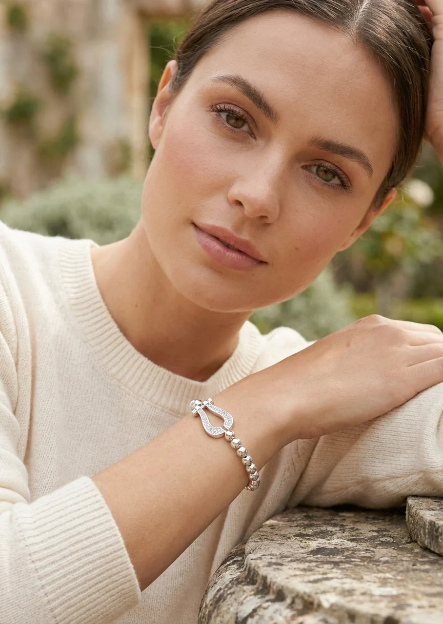 Model wearing a cream jumper with an Envy Silver beaded Bracelet with diamanté detail pendant leaning against a marble wall in a garden setting.