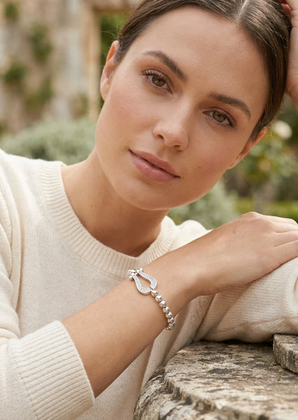 Model wearing a cream jumper with an Envy Silver beaded Bracelet with diamanté detail pendant leaning against a marble wall in a garden setting.