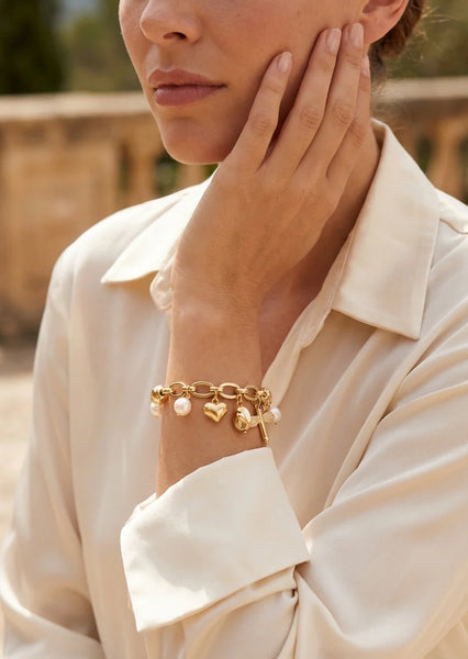 Model wearing a silk cream blouse with an Envy gold beaded and link charm bracelet. Against a garden backdrop.
