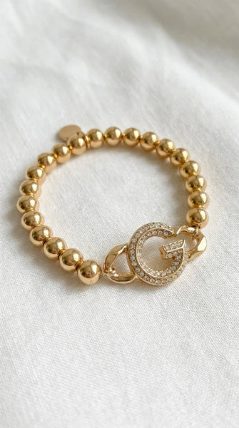 Envy gold beaded elasticated bracelet with G diamanté design pendant. On a white material background.