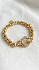 Envy gold beaded elasticated bracelet with G diamanté design pendant. On a white material background.