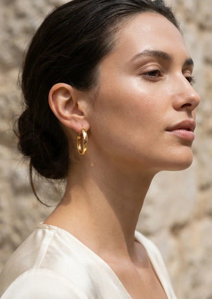 Woman wearing Envy gold hoop earrings with a stone wall background