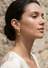 Woman wearing Envy gold hoop earrings with a stone wall background