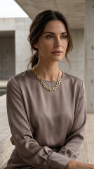 A woman wearing a taupe satin long-sleeve top and an Envy Jewellery chunky gold chain necklace, seated in a modern concrete structure.
