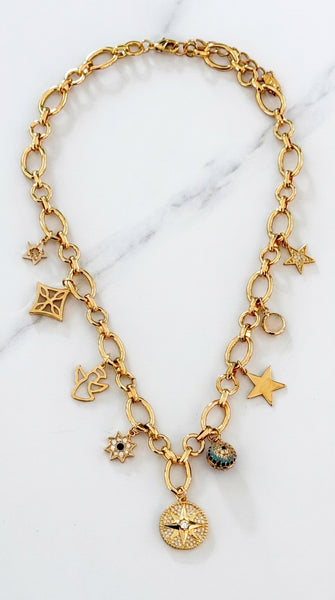 Envy Jewellery gold-tone chain necklace with multiple dangling charms, including stars, a compass, a flower, and small enamel details, displayed on a light marble surface.
