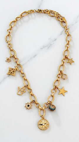 Envy Jewellery gold-tone chain necklace with multiple dangling charms, including stars, a compass, a flower, and small enamel details, displayed on a light marble surface.