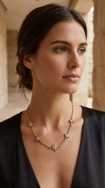 A woman wearing an Envy Jewellery delicate gold necklace with small starburst or flower-like charms, photographed in a softly lit architectural setting. 
