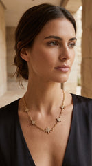 A woman wearing an Envy Jewellery delicate gold necklace with small starburst or flower-like charms, photographed in a softly lit architectural setting. 