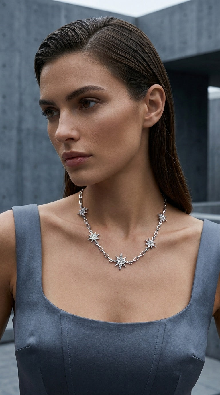 A woman wearing an Envy Jewellery silver-toned necklace with starburst or snowflake-like motifs, photographed in a modern outdoor setting with a gray concrete wall behind her.