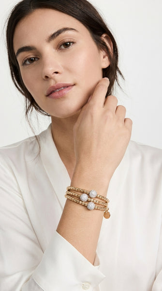 A woman wearing a stack of Envy Jewellery three gold bracelets with white pearl-like beads and small gold charms, paired with a white blouse against a light background.