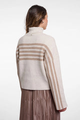 A woman shown from the back wearing a cropped ribbed knit turtleneck sweater in cream and taupe with horizontal stripes across the shoulders and upper back, paired with a pleated taupe skirt, against a plain white studio background.