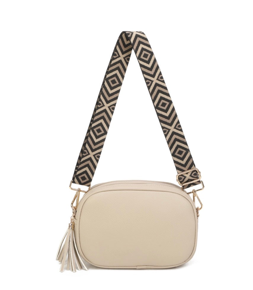 A small crossbody shoulder bag in light beige with a wide, patterned black and beige strap featuring a geometric diamond design, and a matching tassel zipper pull, shown on a plain white background.