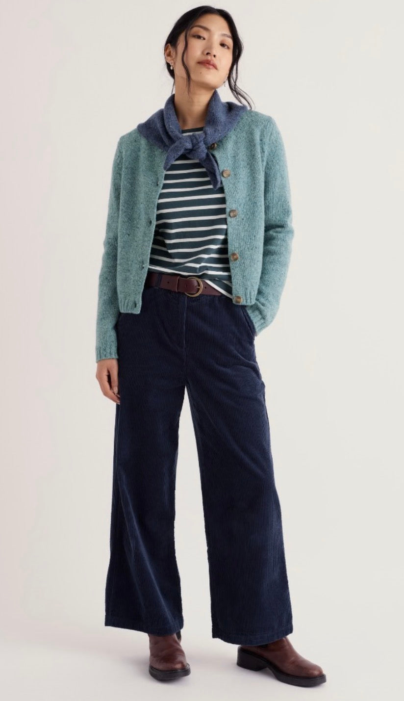 A model stands against a plain light background wearing a light blue-green knit cardigan over a navy and white striped shirt, paired with dark navy wide-leg trousers and brown leather shoes.