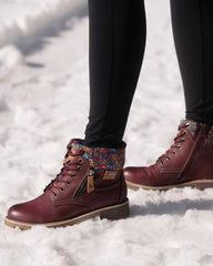 A person wearing burgundy lace-up ankle boots with a colorful knit cuff and black leggings, standing in snow.
